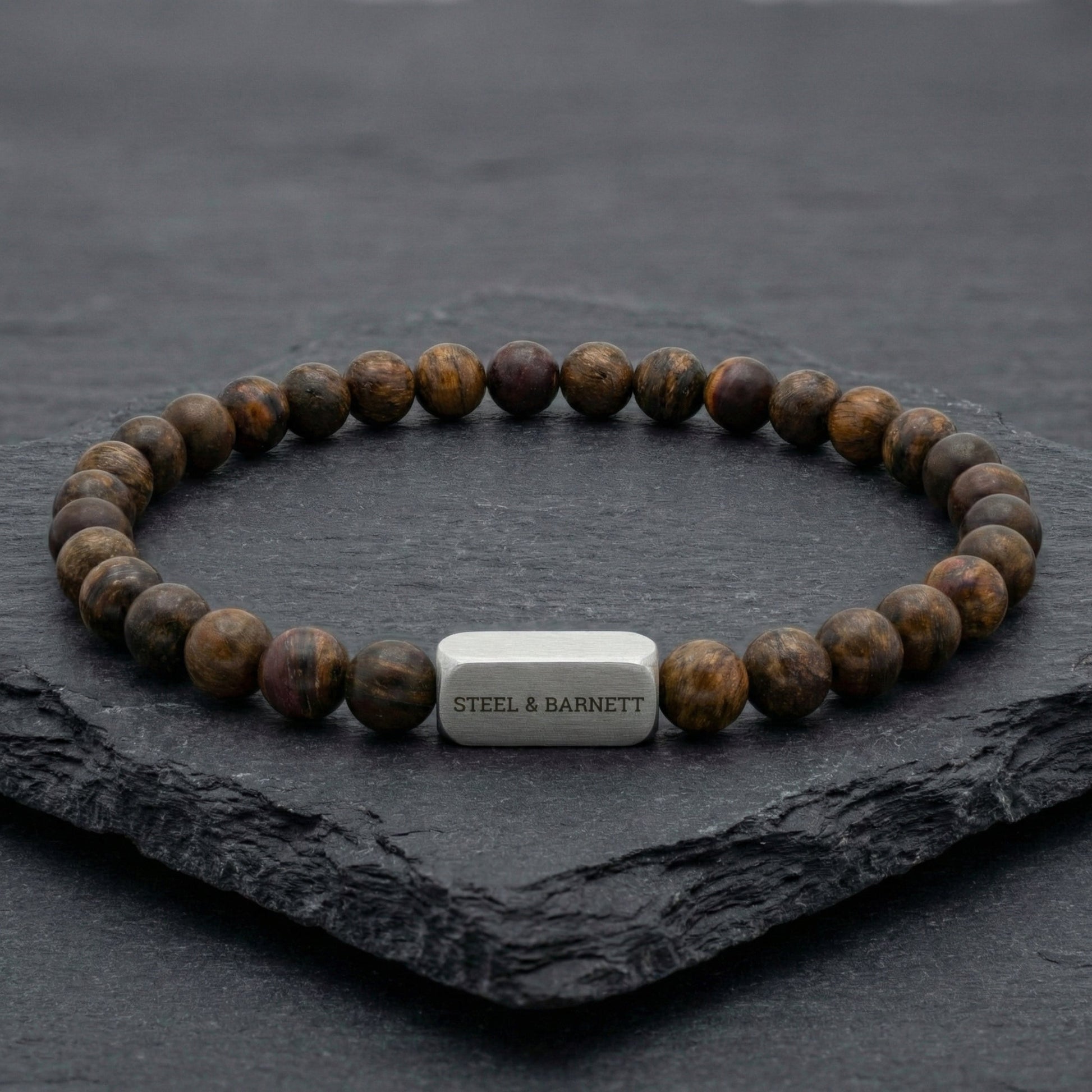 Bronzite beaded men’s bracelet with round brown beads and brushed stainless steel bar on dark stone surface